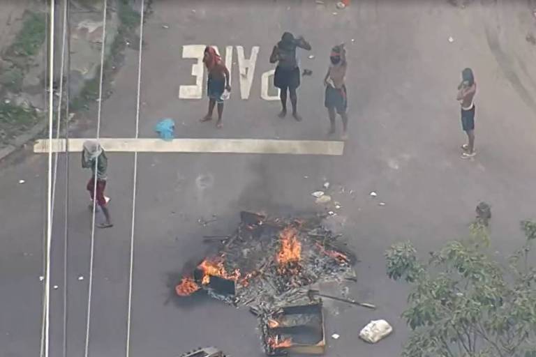 A imagem mostra uma rua com um grupo de pessoas em pé， algumas delas em frente a uma fogueira. Há uma barricada feita de objetos queimados no centro da rua. O ambiente parece tenso， com fumaça visível. Ao fundo， há vegetação e uma linha de postes de eletricidade.