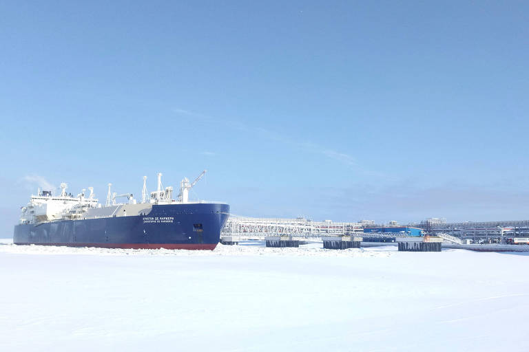 A imagem mostra um grande navio de carga ancorado em uma área coberta de gelo， com um céu claro e azul ao fundo. O navio é de cor azul e possui várias estruturas na parte superior. Há também pequenas embarcações próximas ao navio， todas cercadas por uma vasta extensão de gelo.