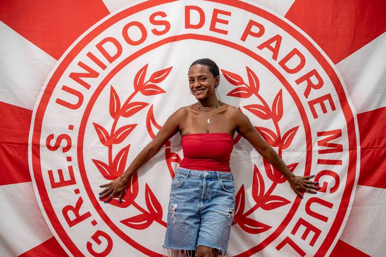 Em foto colorida, mulher de blusa vermelha e bermuda jeans posa em frente a bandeira de uma escola de samba