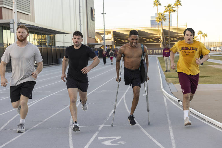 Quatro homens estão correndo em uma pista de atletismo. Dois deles estão em roupas de treino, um deles sem camisa e usando muletas, enquanto os outros dois estão vestidos com camisetas e shorts. Ao fundo, há mais pessoas correndo e palmeiras visíveis ao lado da pista. O ambiente parece ser um centro esportivo.