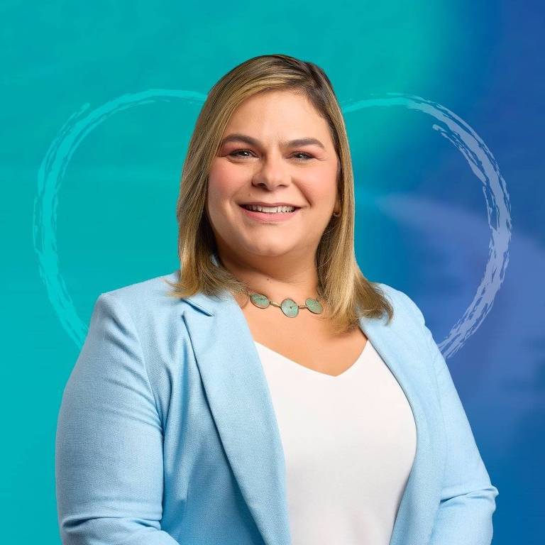 A imagem mostra uma mulher sorridente com cabelo liso e loiro, vestindo um blazer azul claro e uma blusa branca. Ela está posando em frente a um fundo em tons de azul e verde, com um contorno circular desenhado ao fundo. A mulher usa um colar com pedras em tons de verde.