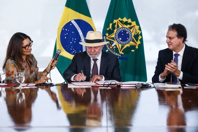 A imagem mostra uma reunião oficial com três pessoas sentadas à mesa. À esquerda， Janja， uma mulher com óculos e cabelo solto segura um celular， observando. No centro， um Lula， com um chapéu branco， assina documentos， enquanto à direita， Camilo Santana aplaude. Ao fundo， estão as bandeiras do Brasil e um símbolo do governo.