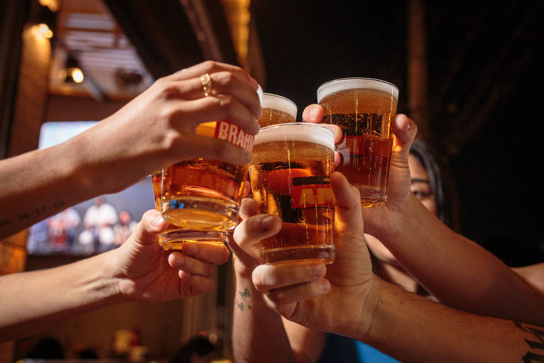 Cinco copos de cerveja estão sendo erguidos em um brinde. As mãos de várias pessoas seguram os copos， que contêm cerveja clara com espuma. Ao fundo， há uma tela com imagens desfocadas e um ambiente iluminado.