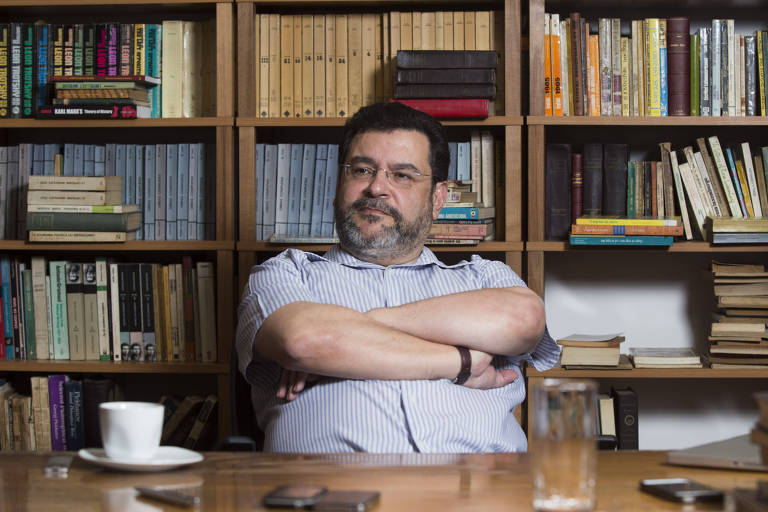 Um homem com barba e óculos está sentado à mesa， com os braços cruzados， olhando para frente. Ao fundo， há uma estante cheia de livros organizados. Na mesa， há uma xícara branca， um copo de água e um celular. O ambiente parece ser um local de trabalho ou uma biblioteca.
