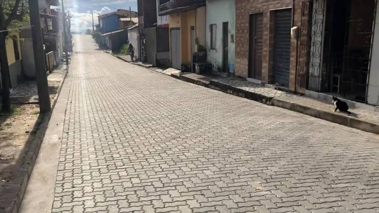 A imagem mostra uma rua pavimentada com pedras, em um dia ensolarado. À esquerda, há algumas árvores e, à direita, casas com portas e janelas visíveis. A rua se estende em linha reta, com uma leve inclinação ao fundo. Não há pessoas visíveis na cena.