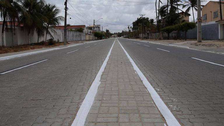 A imagem mostra uma rua vazia, com calçamento em pedras. A via é larga, com faixas brancas no centro, e cercada por muros e algumas árvores ao fundo. O céu está nublado, sugerindo um dia sem sol.