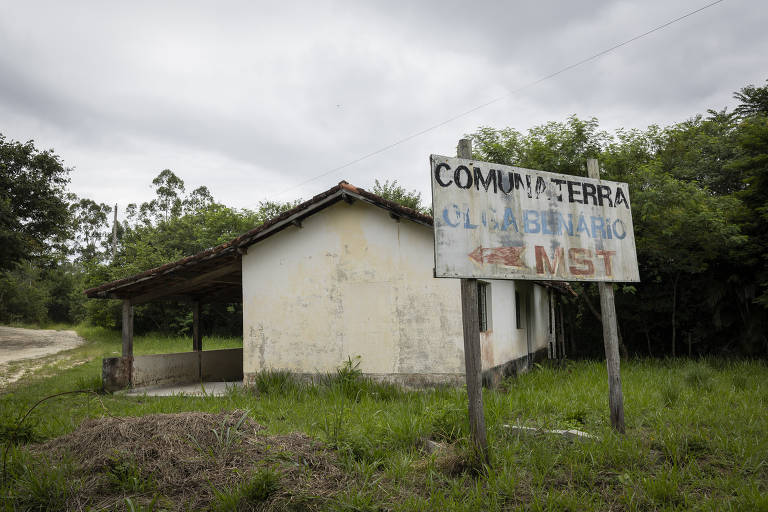 A imagem mostra uma casa simples e abandonada, com paredes brancas e um telhado de duas águas. Na frente da casa, há uma placa de madeira que diz 'COMUNATERRA OLGA BENÁRIO MST'. O ambiente é cercado por vegetação e árvores, e o céu está nublado.