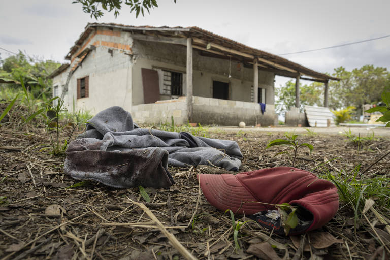 A imagem mostra uma casa abandonada com paredes de alvenaria e janelas com persianas. No primeiro plano， há roupas jogadas no chão， incluindo uma camiseta escura e um boné vermelho. O ambiente é cercado por vegetação e o céu está nublado.