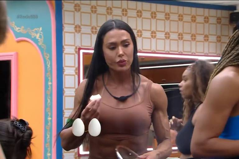 A imagem mostra um grupo de pessoas em um ambiente de reality show. Uma mulher de cabelo longo e liso, vestindo uma blusa marrom, está segurando um pedaço de alimento e parece estar conversando. Ao fundo, outras mulheres estão interagindo, uma delas com cabelo trançado e usando uma blusa azul. O ambiente é decorado com cores vibrantes e possui espelhos nas paredes.