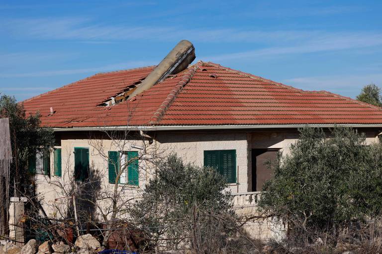 A imagem mostra uma casa com o telhado danificado, onde uma parte do telhado está caída. A casa tem paredes de cor clara e janelas verdes. Ao redor, há algumas árvores e arbustos, e o céu está limpo com algumas nuvens.