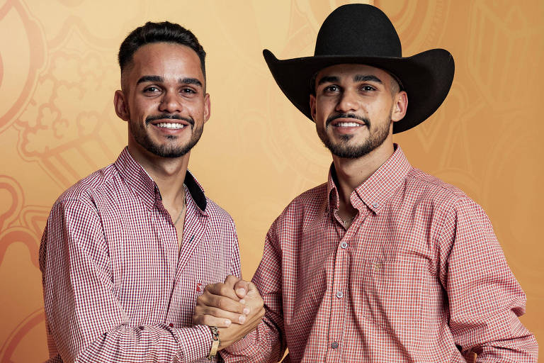 A imagem mostra dois homens sorrindo, ambos vestindo camisas xadrez vermelhas e jeans. O homem à esquerda tem um cabelo curto e usa um cinto com uma fivela decorativa. O homem à direita usa um chapéu preto e está sentado em um banco amarelo, com a mão apoiada na perna. O fundo é de cor laranja com padrões gráficos.