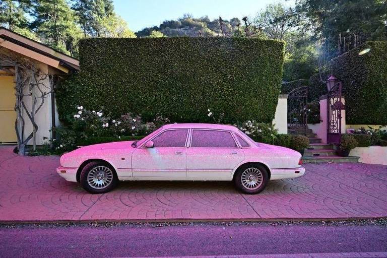 A imagem mostra um carro branco estacionado em uma calçada em frente a uma casa. O carro tem rodas pretas e um design clássico. Ao fundo， há um muro verde bem cuidado e uma entrada com escadas. A área ao redor é arborizada， com plantas e flores visíveis. O piso da calçada é de pedra， com um padrão visível.