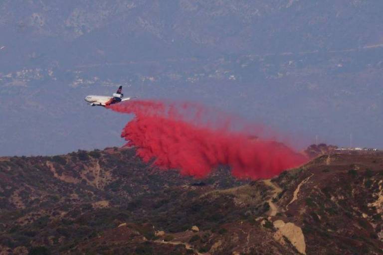 Um avião está voando sobre uma área montanhosa， liberando uma nuvem de tinta vermelha. O fundo apresenta uma paisagem de montanhas e um céu levemente nublado， com uma leve neblina visível.