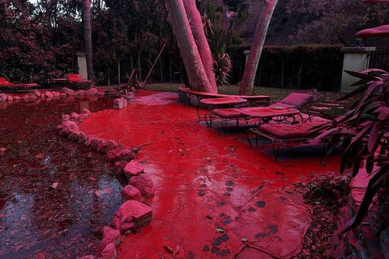 A imagem mostra um espaço externo com um caminho de pedras e uma área de água que reflete uma coloração vermelha. Há espreguiçadeiras dispostas ao redor， algumas cobertas por folhas. O ambiente é cercado por árvores e arbustos， criando uma atmosfera densa e sombria.