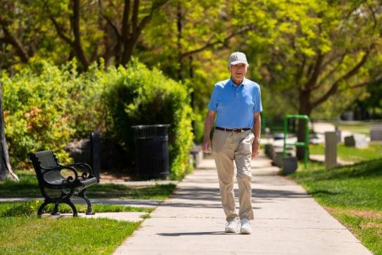 A imagem mostra um homem idoso caminhando em um parque em um dia ensolarado. Ele está vestindo uma camisa azul clara， calças bege e um boné branco. O parque é arborizado， com árvores e arbustos verdes ao fundo， e há um banco preto à esquerda do caminho pavimentado.