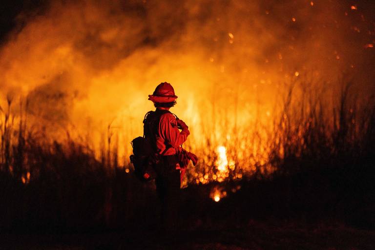 A imagem mostra um bombeiro de costas， vestindo um uniforme laranja e um capacete， observando um incêndio florestal. Chamas intensas e fumaça densa estão visíveis ao fundo， iluminando a cena com uma luz laranja. A vegetação ao redor é alta e seca， e o ambiente parece estar em uma situação crítica devido ao fogo.