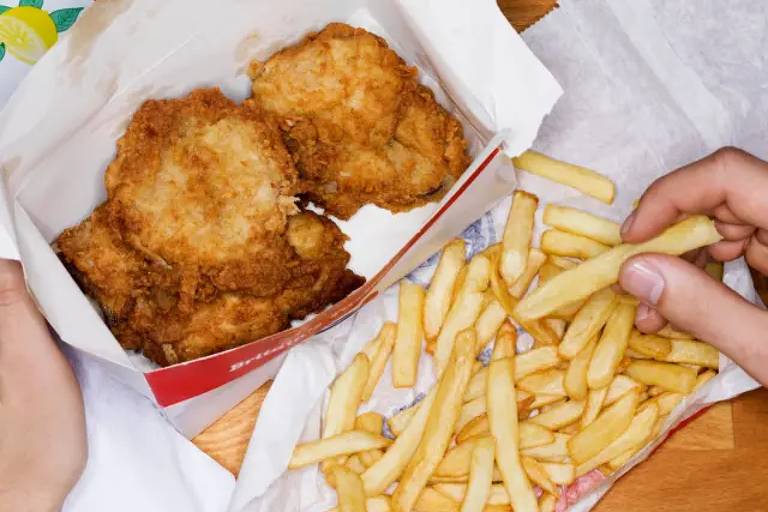 A imagem mostra uma caixa de papel com dois pedaços de frango frito crocante. Ao lado, há uma porção de batatas fritas em um papel. Uma mão está segurando uma batata frita, enquanto a outra mão está segurando a caixa de frango.