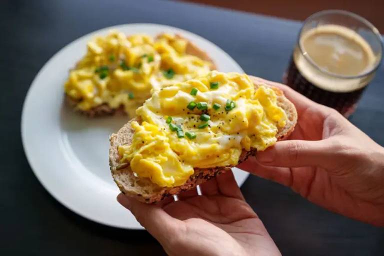 A imagem mostra uma mão segurando uma fatia de pão integral coberta com ovos mexidos, decorados com cebolinha picada. Ao fundo, há um prato com mais uma fatia de pão semelhante e um copo com uma bebida escura, possivelmente café.