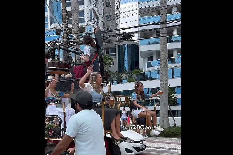 A imagem mostra um grupo de pessoas em um carro aberto， possivelmente durante um desfile ou evento festivo. Algumas pessoas estão acenando e sorrindo， enquanto outras estão sentadas. Ao fundo， há prédios altos e uma vegetação ao redor. A cena é ensolarada e vibrante.