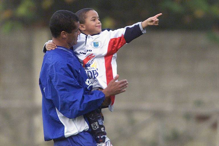 Durante treino da seleção brasileira em Vigo， na Espanha， em 1999， o jogador Rivaldo segura no colo Rivaldinho， seu filho， que aponta para a frente com o dedo indicador da mão esquerda