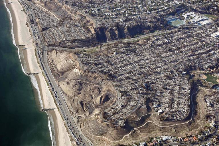 Imagem aérea mostrando uma área costeira com uma praia à esquerda e uma zona urbana à direita. A praia é de areia clara e o mar é de um tom verde-azulado. À direita， há uma área residencial com várias casas dispostas em ruas. A parte superior da imagem apresenta uma área com vegetação e algumas construções. A estrada costeira é visível， com veículos se deslocando.