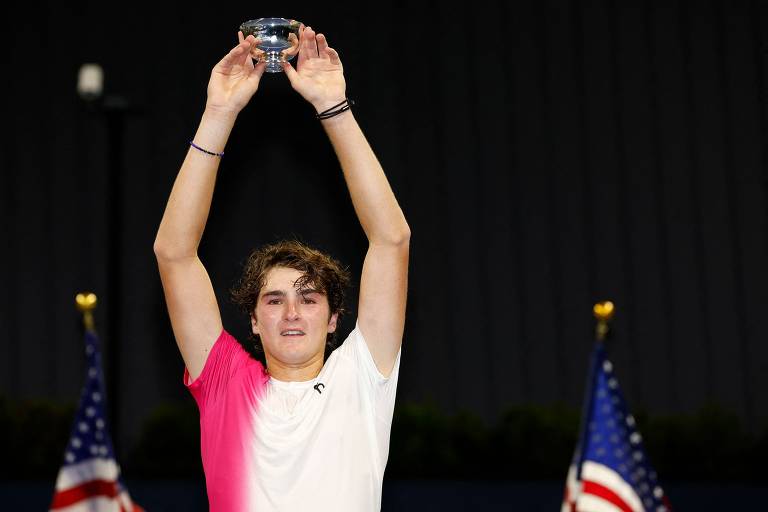 Fotografia mostra o jogador de tênis João Fonseca levantando um troféu acima da cabeça, com uma expressão de felicidade. Ele usa uma camiseta branca com detalhes em rosa. Ao fundo, há bandeiras dos Estados Unidos.