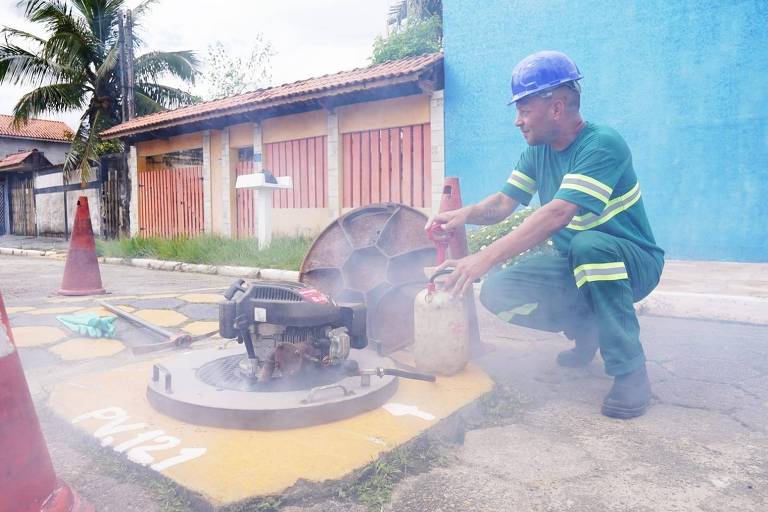 Um trabalhador está próximo a uma tampa de bueiro， levantando-a enquanto fumaça sai de dentro. Ele usa um capacete azul e um uniforme verde com listras. Ao fundo， há casas com paredes coloridas e uma árvore. O ambiente parece ser urbano.