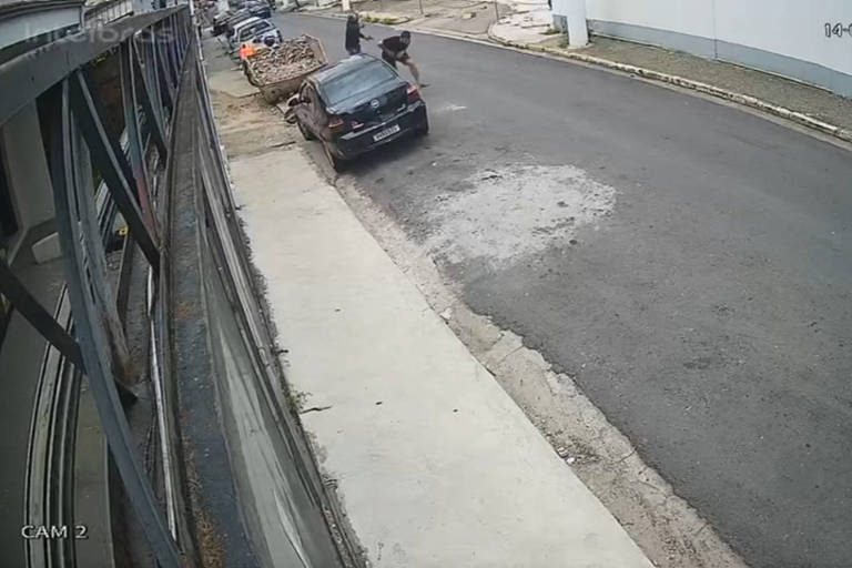 A imagem mostra uma cena de rua capturada por uma câmera de segurança. À esquerda， há uma estrutura que parece ser um prédio ou uma parede. No centro， um carro preto está estacionado na calçada， enquanto uma pessoa está próxima a ele. Ao fundo， é possível ver uma rua pavimentada e uma esquina. Há um carro estacionado do lado esquerdo da imagem， perto da calçada esquerda. Um homem parece se agachar enquanto corre e， atrás dele， há outro homem com capacete de moto， a pouco mais de um metro.