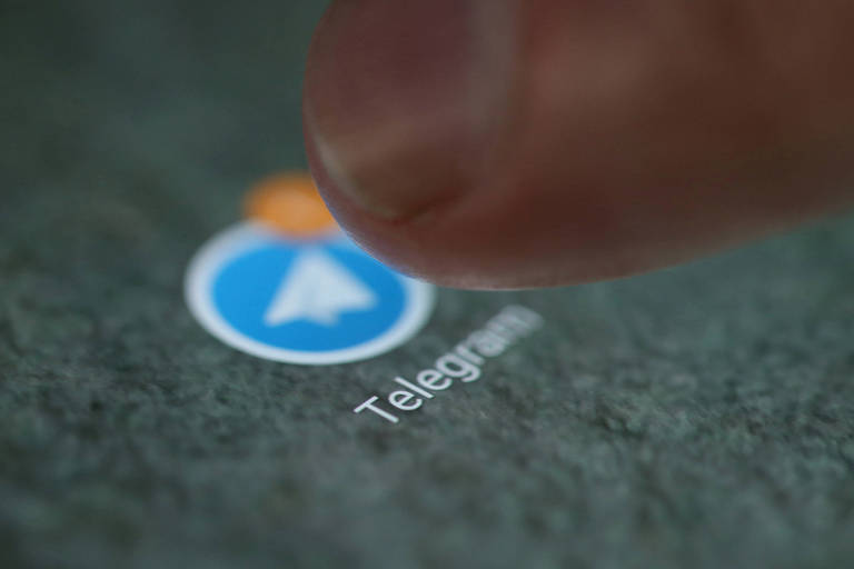 A imagem mostra um dedo prestes a tocar o ícone do aplicativo Telegram， que é representado por um círculo azul com um símbolo de avião de papel branco. Abaixo do ícone， está escrito 039;Telegram039;. O fundo da imagem é de uma superfície escura