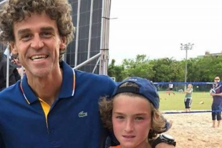 A imagem mostra um homem e um menino sorrindo para a câmera. O homem tem cabelo cacheado e usa uma camisa azul com detalhes em amarelo. O menino， que está ao lado dele， usa uma camiseta laranja listrada e um boné azul. Ao fundo， há um campo com pessoas e equipamentos esportivos.