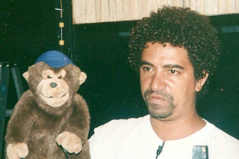 Um homem com cabelo cacheado e barba segura um boneco de macaco que usa um chapéu azul. O fundo é escuro， com uma estrutura que parece ser um palco ou cenário. O homem tem uma expressão séria.