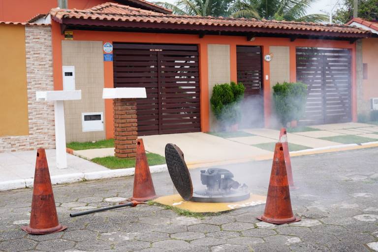 A imagem mostra uma rua residencial com uma tampa de bueiro levantada, de onde sai fumaça. Ao redor da tampa, há cones de sinalização laranja. Ao fundo, pode-se ver casas com fachadas coloridas e portões. A cena sugere uma atividade de manutenção ou reparo na via.