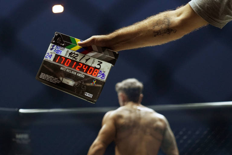 A imagem mostra uma mão segurando uma claquete de filmagem, com informações escritas à mão. Ao fundo, um lutador de costas, com um corpo musculoso e tatuagens visíveis, está se preparando para uma luta em um octógono. A iluminação é baixa, com um foco de luz no centro da cena.