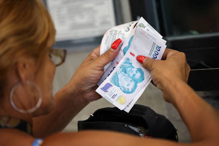 Uma mulher com cabelo loiro e ondulado está segurando notas de euro， contando-as. Ela usa anéis e tem unhas pintadas de vermelho. Ao fundo， há um caixa eletrônico.