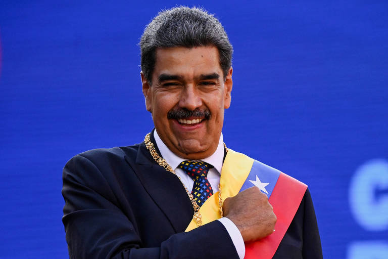 Um homem sorridente está posando com a mão no peito， usando um terno escuro e uma faixa colorida que representa a bandeira da Venezuela. O fundo é azul， e ele parece estar em um evento formal