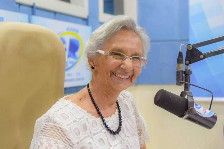 A imagem mostra uma mulher idosa sorrindo， sentada em um estúdio de rádio. Ela usa óculos e tem cabelo grisalho preso. A mulher está vestida com uma blusa branca e um colar escuro. Ao fundo， há um microfone e uma parede com elementos de estúdio， incluindo um logotipo visível.