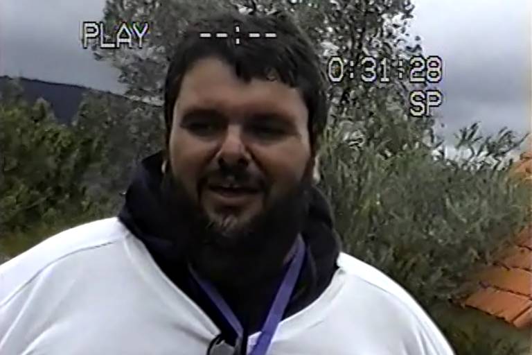 Um homem com barba e cabelo escuro， vestindo uma camiseta branca e um moletom preto， está sorrindo e falando. Ao fundo， há vegetação verde e um céu nublado. A imagem tem uma sobreposição de texto que indica 039;PLAY039; e um temporizador que marca 0:31:28.   