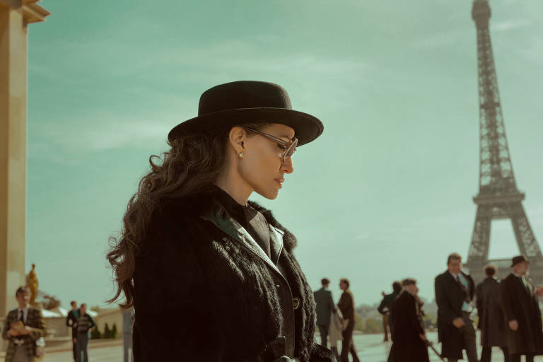 A imagem mostra uma mulher com cabelo cacheado e um chapéu preto, usando óculos de sol e um casaco escuro. Ela está em um local ao ar livre em Paris, com a Torre Eiffel visível ao fundo. Há várias pessoas ao redor, algumas em trajes formais, e o céu tem uma tonalidade esverdeada.