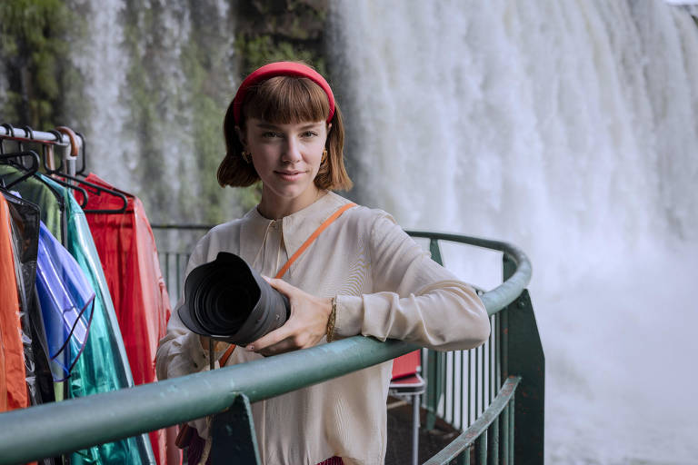 A imagem mostra uma mulher segurando uma câmera em um local próximo a uma cachoeira. Ela está usando uma blusa clara e um lenço vermelho na cabeça. Ao fundo, há uma cachoeira e, à sua esquerda, um cabide com roupas coloridas, incluindo capas de chuva em várias cores. O ambiente é úmido e natural, com vegetação ao fundo.