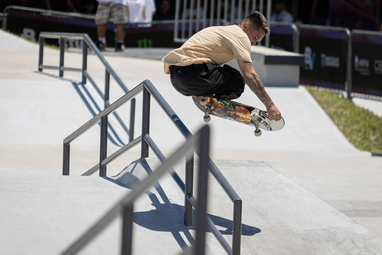 Um skatista está realizando um salto sobre uma barra de ferro em um parque de skate. Ele está vestido com uma camiseta clara e calças escuras, enquanto segura seu skate com uma das mãos. O ambiente é ensolarado, com outras pessoas visíveis ao fundo.