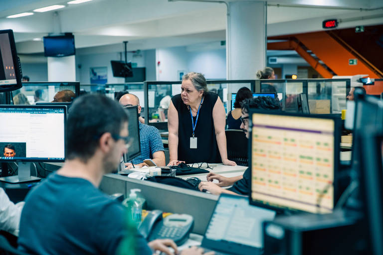 A imagem mostra uma redação de jornal com várias pessoas trabalhando em estações de trabalho. No primeiro plano， um homem de camiseta escura está concentrado em seu computador， enquanto uma mulher de cabelo grisalho e blusa preta está em pé， interagindo com os colegas. Ao fundo， há várias telas de computador exibindo informações e gráficos. O ambiente é bem iluminado e organizado， com divisórias entre as mesas.