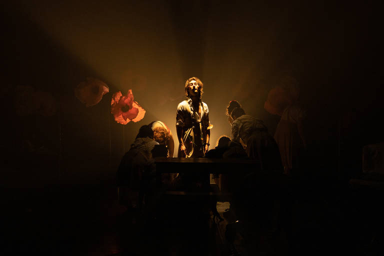 A imagem mostra uma cena de teatro com um foco de luz iluminando um ator em pé no centro, cercado por outros atores que estão agachados ao seu redor. O fundo é escuro, com flores grandes e coloridas visíveis, criando um ambiente dramático. A iluminação destaca a expressão do ator principal, enquanto os outros parecem estar em uma posição de adoração ou atenção.