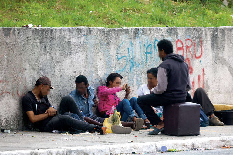 A imagem mostra um grupo de cinco pessoas sentadas em um espaço urbano， encostadas em uma parede de concreto. Algumas pessoas estão interagindo entre si， enquanto outras parecem estar distraídas. O ambiente é simples， com grama ao fundo e uma calçada à frente. Há grafites visíveis na parede， e um dos indivíduos está sentado em um banco ou caixa.