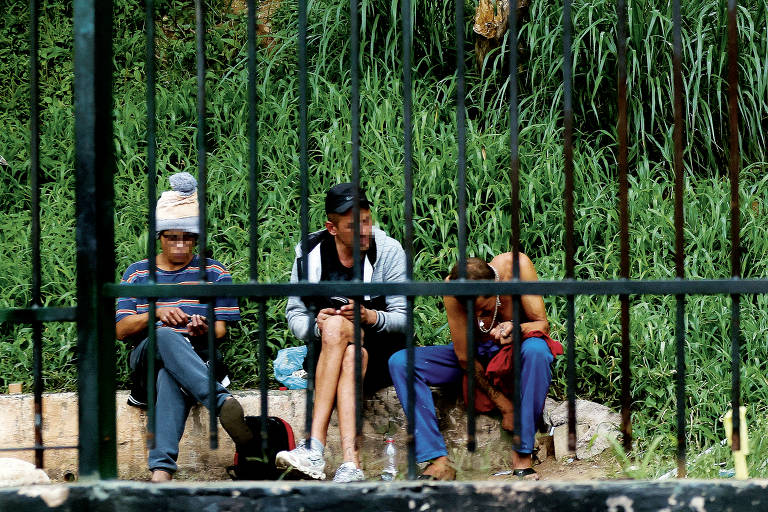 A imagem mostra três pessoas sentadas em um banco de madeira, com uma grade de metal ao fundo. As pessoas estão em um ambiente com grama alta. A primeira pessoa, à esquerda, usa um gorro e uma camiseta listrada. A segunda pessoa, no centro, está vestindo uma camiseta preta e calças claras. A terceira pessoa, à direita, está sem camisa e usa calças coloridas. Todas parecem estar em um momento de descanso.