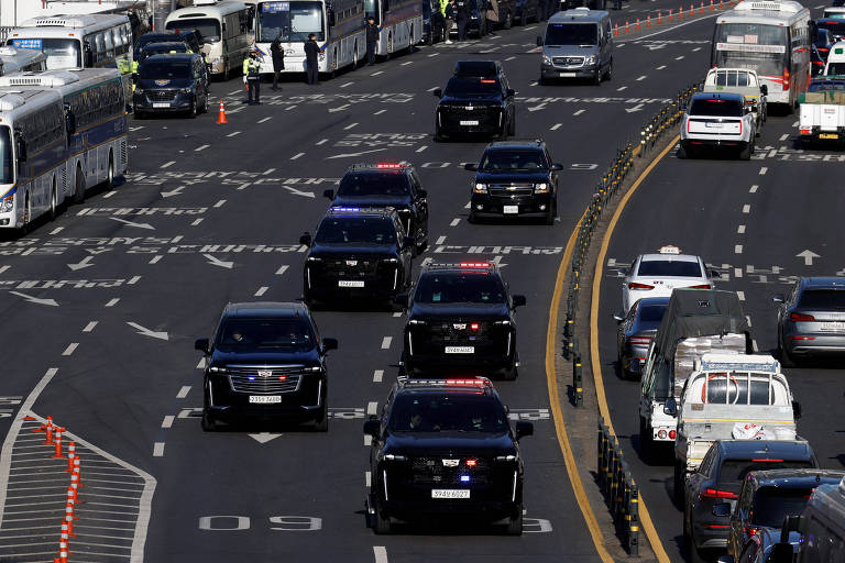 A imagem mostra uma via movimentada com vários veículos， incluindo carros pretos que parecem ser de autoridades， em um tráfego intenso. Há ônibus e outros carros em diferentes faixas， com algumas marcas de sinalização no chão