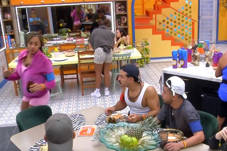 A imagem mostra uma cozinha de um reality show, onde várias pessoas estão interagindo. À mesa, há três homens e uma mulher, com pratos de comida e uma tigela com maçãs verdes. Ao fundo, algumas pessoas estão preparando alimentos e outras estão em pé, conversando. O ambiente é colorido, com paredes amarelas e detalhes em azul. Há uma escada ao fundo e várias garrafas e utensílios na bancada.