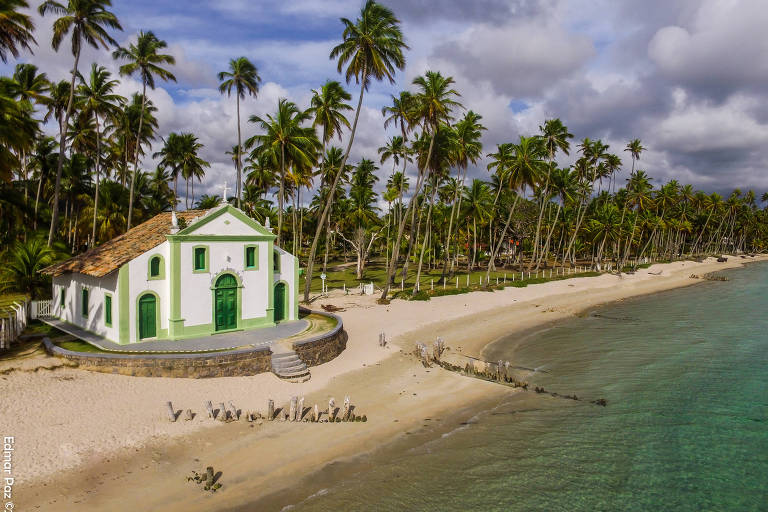 Imagem aérea mostra praia vazia， com areia bem clara e mar esverdeado. A paisagem， paradisíaca， é composta ainda por coqueiros que formam uma linha à beira do areal， e com uma capela， cor branca， em estilo antigo de arquitetura