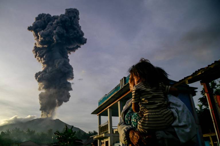 A imagem mostra uma erupção vulcânica com uma grande coluna de fumaça subindo ao céu. Em primeiro plano, uma criança está sentada nas costas de um adulto, ambos observando a erupção. O cenário inclui uma construção ao fundo, que parece ser uma casa ou um abrigo, e vegetação ao redor.