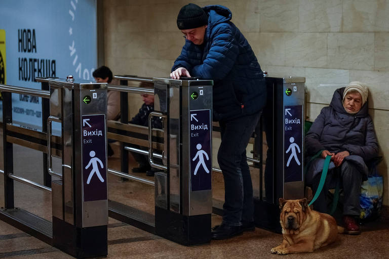 Ao lado de uma catraca de metrô， um homem com casaco se apoia e uma mulher senta junto à parede， com seu cachorro marrom na coleira à sua frente