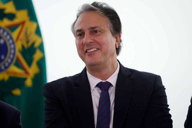 Um homem com cabelo liso e levemente ondulado， vestindo um terno escuro e uma gravata listrada， está sorrindo. Ao fundo， há uma bandeira do Brasil， com as cores verde， amarelo e azul. O ambiente parece ser um evento formal.